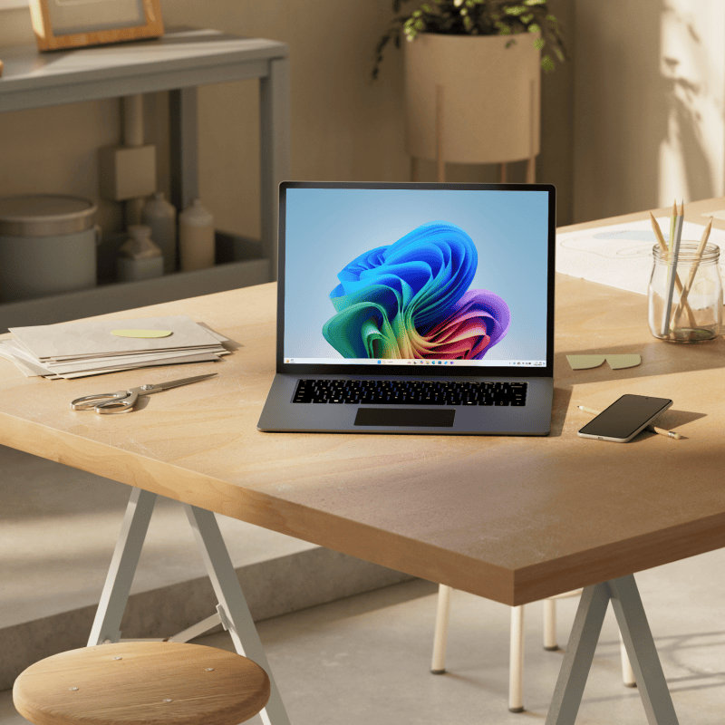 An open laptop with the rainbow bloom screen sitting on a table with office supplies, shelves, and a plant in the background