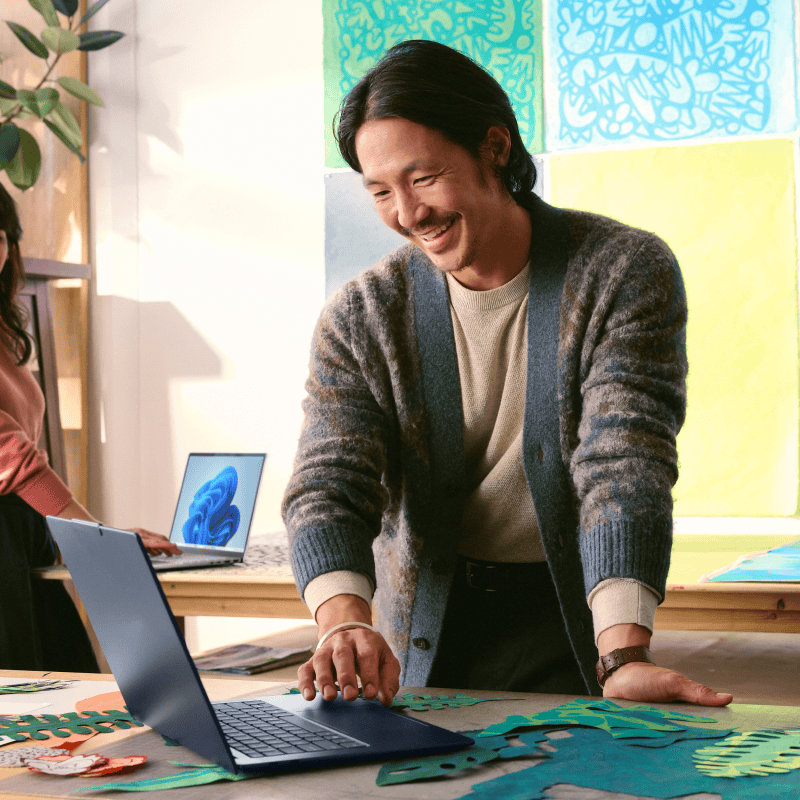 A person smiling and standing at a table with an open laptop surrounded by art