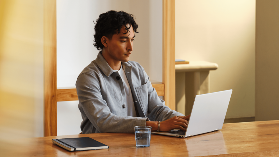 A man sitting at a wooden table, working on a laptop. A closed notebook and a glass of water are also on the table, with wooden furniture and light-colored walls in the background.
