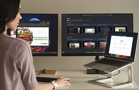 A woman sitting at a desk with three screens: a laptop on a stand and two external monitors. A notepad and pen are also on the desk.