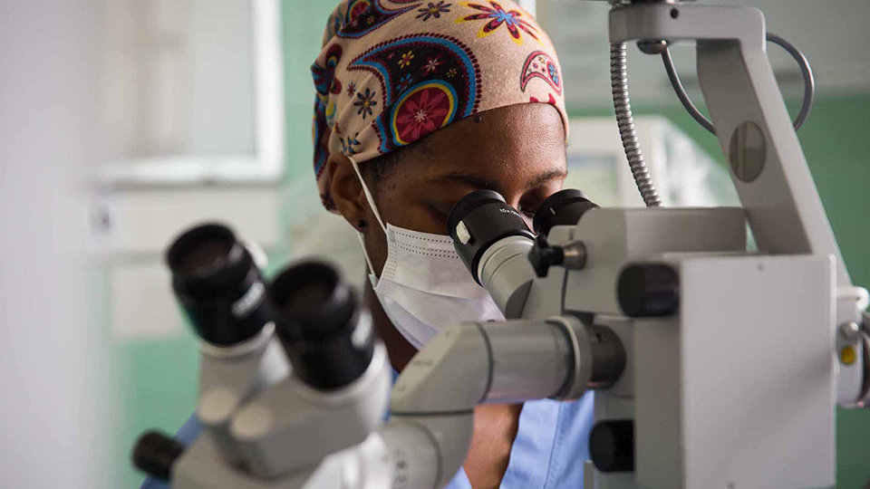 A person wearing a headscarf and medical face mask looks through a microscope